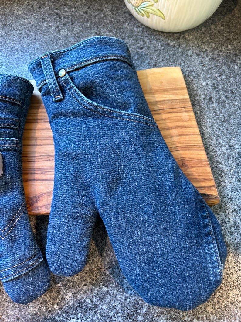 May include: A pair of oven mitts crafted from blue denim fabric, featuring classic jean details such as a curved pocket opening, a metal rivet, and a belt loop at the wrist. The mitts have visible topstitching and a rounded thumb section.