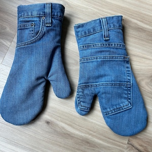 May include: A pair of blue denim oven mitts crafted from repurposed jeans. One mitt features the front waistband, belt loops, and pocket details, while the other displays the back pocket and horizontal stitching patterns of the original denim fabric.