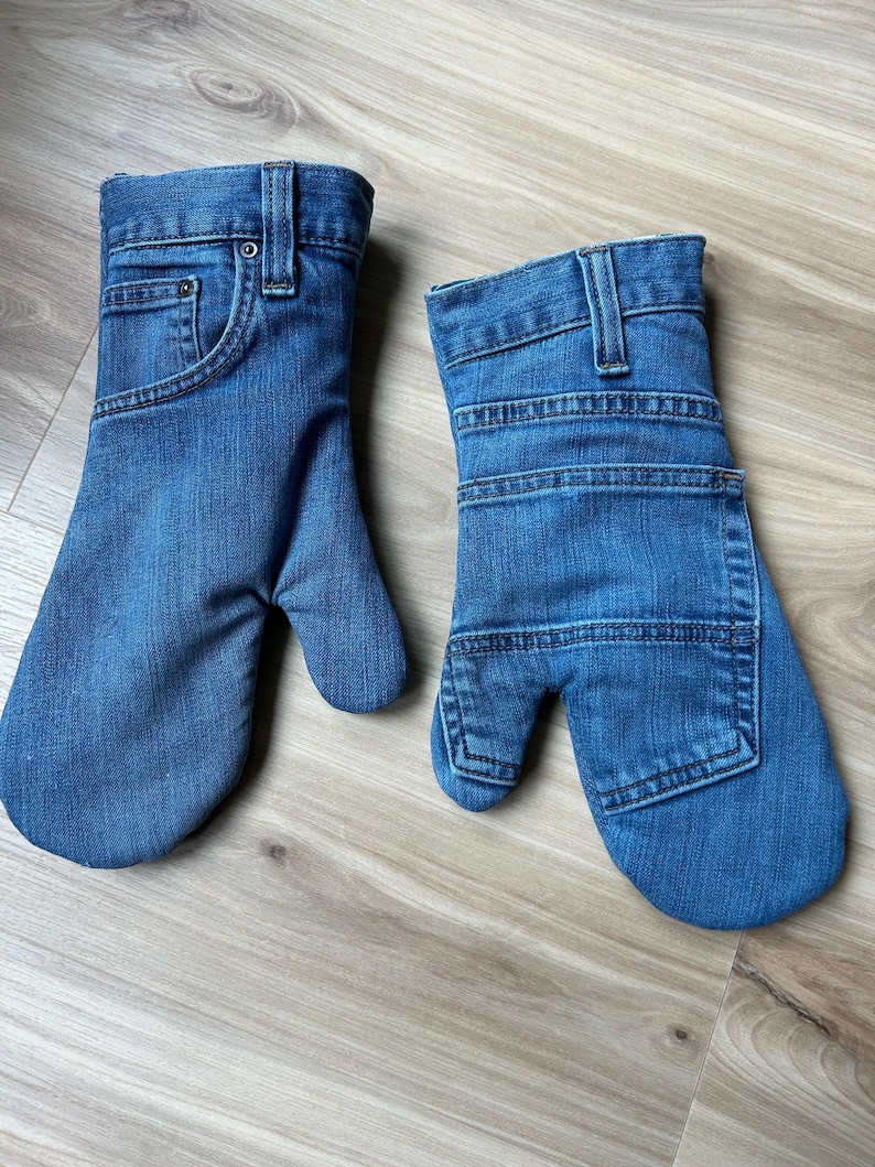May include: A pair of blue denim oven mitts crafted from repurposed jeans. One mitt features the front waistband, belt loops, and pocket details, while the other displays the back pocket and horizontal stitching patterns of the original denim fabric.