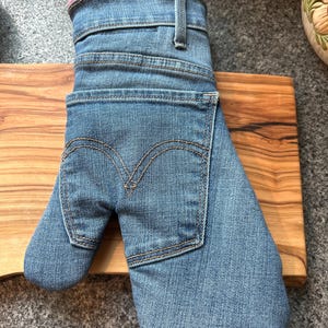 May include: An oven mitt constructed from blue denim fabric, featuring the distinct waistband, belt loop, and back pocket design of a pair of jeans. The pocket displays the characteristic double-arched stitching pattern in gold thread.