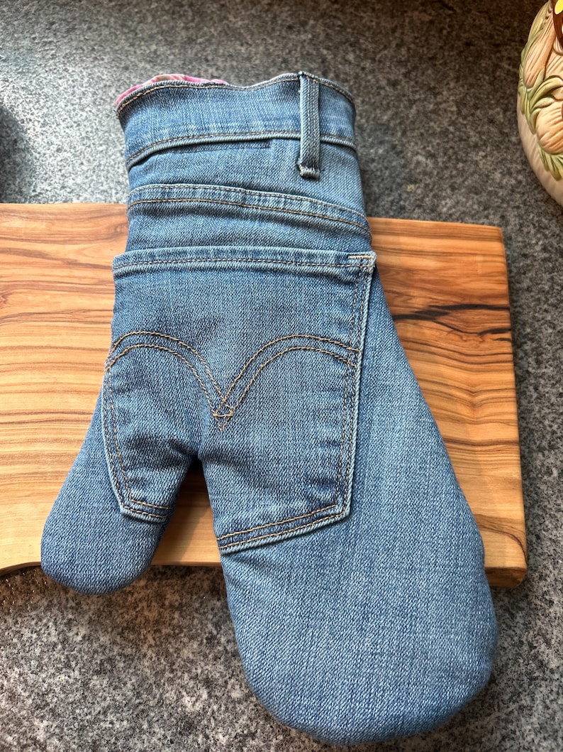May include: An oven mitt constructed from blue denim fabric, featuring the distinct waistband, belt loop, and back pocket design of a pair of jeans. The pocket displays the characteristic double-arched stitching pattern in gold thread.