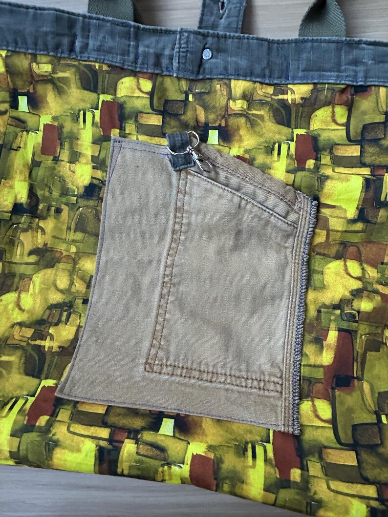 Puede incluir: Bolso tote hecho a mano con un dise&ntilde;o &uacute;nico. El bolso presenta un colorido patr&oacute;n abstracto en tonos amarillos, verdes y marrones. Tiene un bolsillo beige y correas verde oliva. Un llavero plateado est&aacute; sujeto al bolsillo.