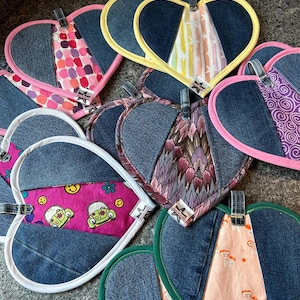 May include: A collection of heart-shaped denim pot holders with various patterned fabric inserts. Each pot holder has a different colored trim, including pink, yellow, and green. They have a small loop for hanging.