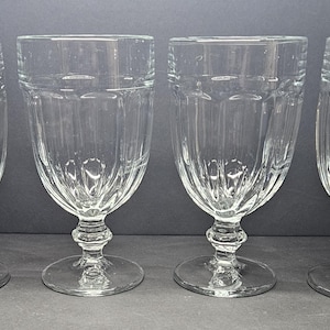 May include: A set of four clear glass goblets featuring a vertical ribbed pattern along the lower bowl and a thick, rounded stem base. Each glass has a smooth, flared rim and a sturdy foot, showcasing a transparent design with light-reflecting facets.