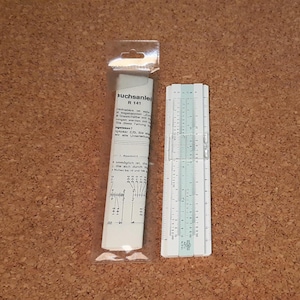 Może przedstawiać: Suwak logarytmiczny i jego opakowanie są pokazane na brązowej powierzchni. Suwak logarytmiczny jest biały z czarnymi i turkusowymi oznaczeniami. Opakowanie to przezroczysta plastikowa torba zawierająca zwinięty papier z tekstem.