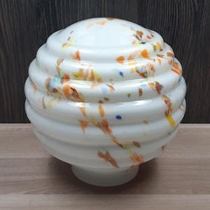 Könnte beinhalten: Weißer, kugelförmiger Lampenschirm aus Glas mit einem wirbelnden, gerippten Design. Er ist mit farbigen Flecken in Orange, Gelb und Blau verziert. Der Lampenschirm hat einen kleinen, runden Sockel.