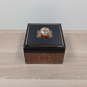 Puede incluir: Una caja decorativa negra con un pomo de cristal en la tapa. La caja tiene diseños dorados ornamentados en la tapa y el frente. La caja está sobre una superficie de madera.