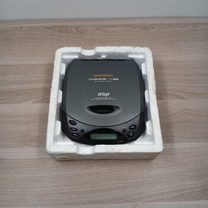 Könnte beinhalten: Grauer tragbarer CD-Player von Daewoo, mit den Schriftzügen "Daewoo" und "asp". Der Player befindet sich in einem weißen Schaumstoffbehälter. Er verfügt über ein digitales Display und Bedientasten.