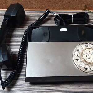 May include: A black rotary phone with a silver faceplate and a black rotary dial. The phone has a black handset and a coiled cord.
