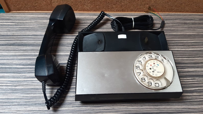 May include: A black rotary phone with a silver faceplate and a black rotary dial. The phone has a black handset and a coiled cord.