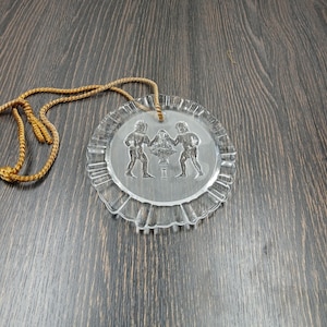 Könnte beinhalten: Ein klarer, kreisförmiger Glasornament mit dekorativem Rand und einer goldfarbenen Kordel. Das Ornament zeigt ein geätztes Design von zwei Figuren, die einen Blumenkorb flankieren. Das Ornament hat einen Durchmesser von etwa 10 cm.