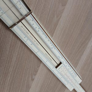 Może przedstawiać: Zabytkowa suwmiarka, z kremowym korpusem i czarnymi i czerwonymi oznaczeniami. Suwak ma metalową ramę i zawiera tekst "MADE IN GERMANY" oraz "A.W. FABER-CASTELL 1/87 RIETZ".