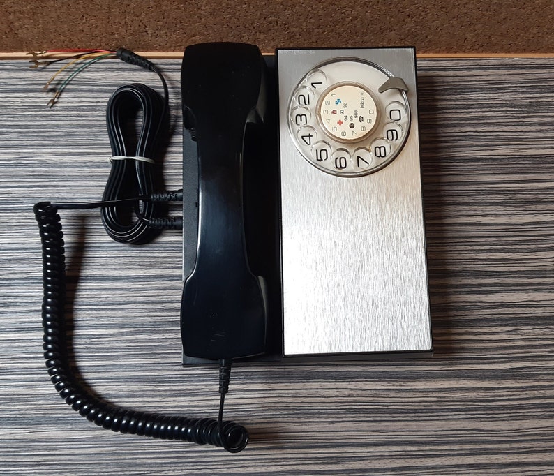 May include: A black rotary dial phone with a silver faceplate and a black coiled cord. The phone has a rotary dial with numbers 1 through 9 and a star and pound symbol. The phone is on a wooden surface.