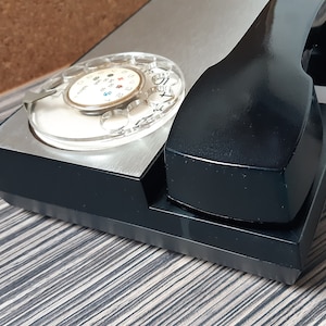May include: A black rotary dial phone with a silver faceplate and a black handset. The phone has a clear dial with black numbers and a black cord.