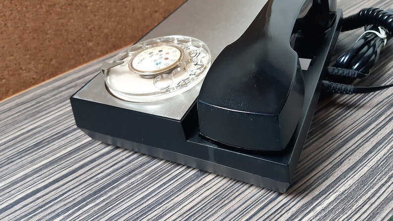 May include: A black rotary dial phone with a silver faceplate and a black handset. The phone has a clear dial with black numbers and a black cord.