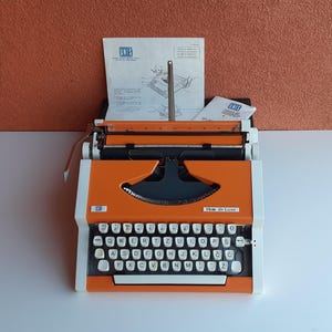 Pode incluir: Máquina de escrever UNIS IBM de Luxe laranja e branca com teclado preto e manual. A máquina de escrever tem um design retro com carro de metal e alimentação de papel. O teclado tem teclas brancas com letras pretas.