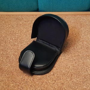 Könnte beinhalten: Schwarzes Leder-Münzfach mit gewölbtem Oberteil und Druckknopfverschluss. Der Innenraum ist mit dunkelblauem Stoff gefüttert. Die Geldbörse ist geöffnet und gibt den Innenraum frei. Die Geldbörse liegt auf einer Korkoberfläche.