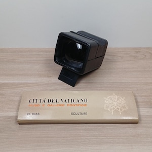 Op de afbeelding: Een zwarte, rechthoekige viewer met een scharnierende standaard staat boven een beige doos. De doos heeft Italiaanse tekst, waaronder "Città del Vaticano" en "24 Dias". De viewer is van plastic.