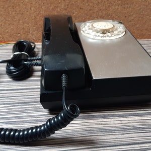 May include: A black rotary phone with a silver faceplate and a coiled black cord. The phone has a black handset and a push-button dial.