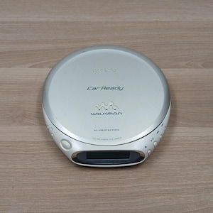 Può includere: Lettore CD Sony Walkman argentato con la scritta "Car Ready" e "G Protection". Il lettore CD portatile ha un piccolo schermo e diversi pulsanti di controllo. Il dispositivo è su una superficie di legno.