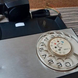 May include: A black rotary phone with a silver faceplate and a clear plastic dial. The dial has numbers 1 through 0 and symbols for international calls. The phone has a black handset and a black cord.