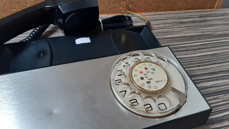 May include: A black rotary phone with a silver faceplate and a clear plastic dial. The dial has numbers 1 through 0 and symbols for international calls. The phone has a black handset and a black cord.