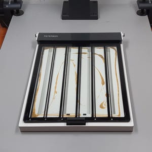 May include: A Paterson film developing tank with a white base, black frame, and clear interior. The interior has a white surface with gold swirls. The rectangular tank sits on a grey surface.