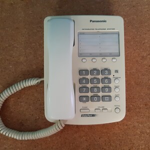 Może przedstawiać: Biały zintegrowany system telefoniczny Panasonic z małym wyświetlaczem LCD, klawiaturą z przyciskami do numerów, liter i funkcji specjalnych oraz białą słuchawką z kablem spiralnym.