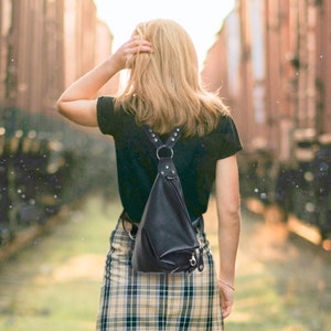 Peut inclure: Un sac à dos en bandoulière en cuir noir avec des détails argentés est porté sur un t-shirt noir et une jupe à carreaux. La jupe est beige et noire. Des baskets blanches complètent la tenue. La personne s'éloigne de la caméra.