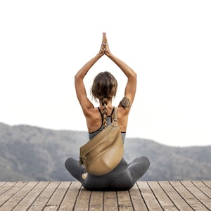 May include: A person in a yoga pose, wearing a gray sports bra and leggings, with a tan leather backpack. The person is sitting on a wooden deck with a mountain range in the background. A heart-shaped charm hangs from the backpack.
