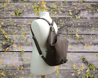 May include: A brown leather backpack purse displayed on a mannequin. The bag has adjustable straps and a single shoulder strap. The background is a wooden surface with leaves.