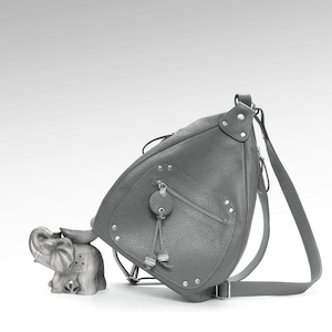 May include: A gray leather sling bag with a textured surface, featuring a zippered pocket and decorative metal studs. The bag has a long adjustable strap and a drawstring closure. An elephant figurine is next to the bag.