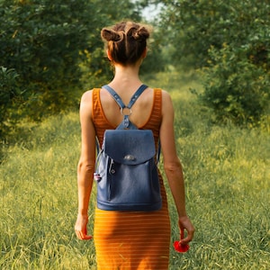 May include: A woman wearing an orange dress and a blue leather backpack walks through a field of tall grass. The backpack has a flap closure and adjustable straps. The woman is holding a red flower.