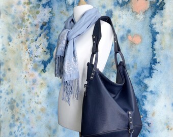 May include: A navy blue leather hobo bag with a textured finish, displayed on a mannequin alongside a light blue paisley scarf with fringe. The bag has a rounded shape and a shoulder strap. The background features a blue and white watercolor design.