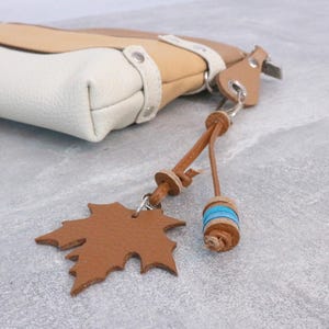 Può includere: Una borsa in pelle tricolore con un ciondolo a forma di foglia d'acero. La borsa presenta sezioni bianche, beige e marroni. Il ciondolo è in pelle marrone e il nappa ha accenti blu.