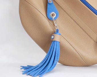 May include: A blue tassel keychain with a light brown leather accent and a silver-tone clasp. The tassel is attached to a blue leather tag with a silver ring. The background is a light brown leather bag.