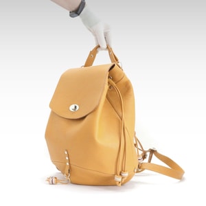 May include: A mustard-yellow leather backpack with a flap closure and silver-tone hardware. The bag has a drawstring closure, adjustable straps, and a top handle. The backpack is held up by a gloved hand.