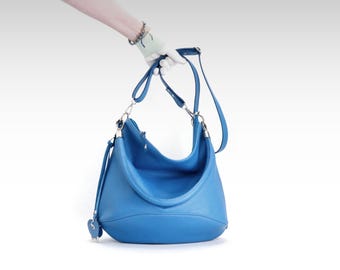 May include: A bright blue leather shoulder bag with silver-tone hardware. The bag has a curved shape, a long adjustable strap, and a small heart-shaped charm. The bag is held by a gloved hand against a white background.