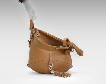 May include: A tan leather hobo bag with a rounded shape and a long shoulder strap. The bag features silver-tone hardware, including rivets and a tassel. The bag is held by a gloved hand against a white background.
