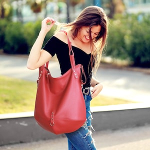 May include: A large, red leather shoulder bag with a rounded shape and two handles. The bag has silver-coloured hardware and a zip closure. The bag is being held by a person wearing a black top and blue jeans.