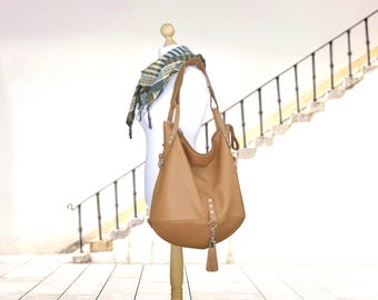 May include: A tan leather shoulder bag with a tassel, displayed on a mannequin. The bag has a rounded shape and a long strap. A scarf with green and blue stripes is draped over the mannequin's shoulder.
