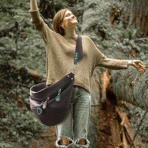 May include: A dark brown leather sling bag with a teal zipper pull and heart charm. The bag has a wide strap with teal accents. The bag is worn across the body. The person in the image is wearing a tan sweater and jeans.