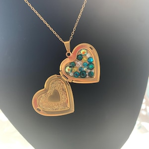 May include: A gold heart-shaped locket necklace with a chain. The locket is open, revealing a heart-shaped compartment filled with green, blue, and yellow crystals.