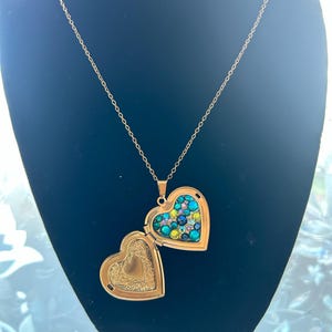 May include: A gold heart-shaped locket necklace with a chain. The locket is open, revealing a heart-shaped interior with a design of green, blue, and clear rhinestones.