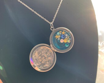 May include: A silver chain necklace with a round locket pendant. The locket is open, revealing a blue resin filled with gold and blue glitter, pearls, and seashells.