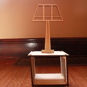 May include: A wooden lamp with a geometric shade sits on a small wooden table with a shelf. The table is made of light wood and has a simple design.