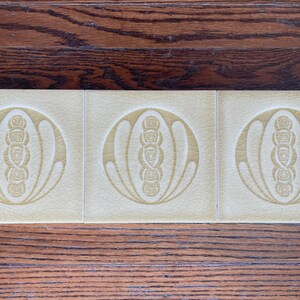 May include: Three cream-colored ceramic tiles with a raised design of a stylized flower with a circle around it. The design is in a light yellow color.