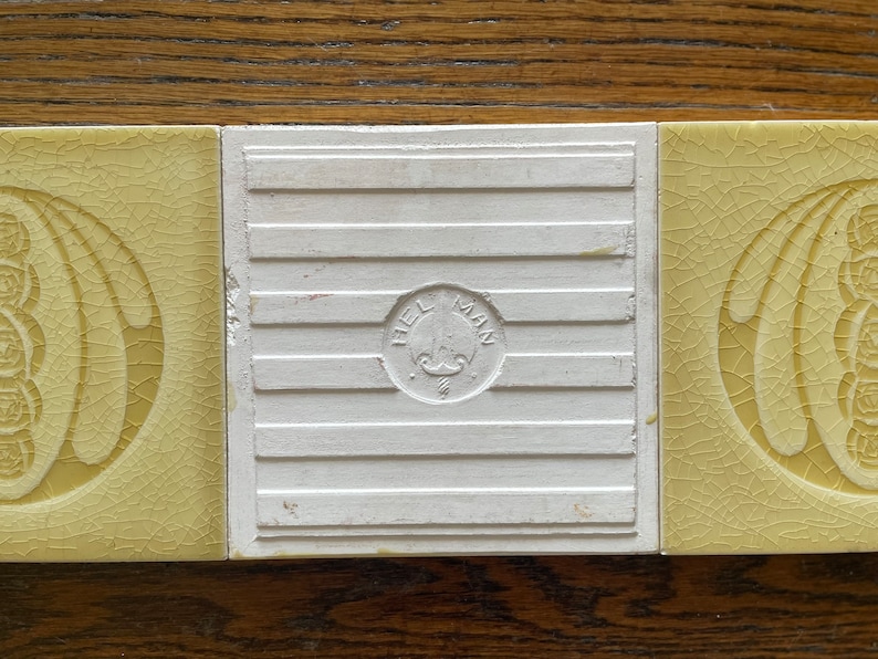 May include: Three yellow ceramic tiles with a decorative floral design. The center tile has a white background and a circular stamp that reads "Hellman".