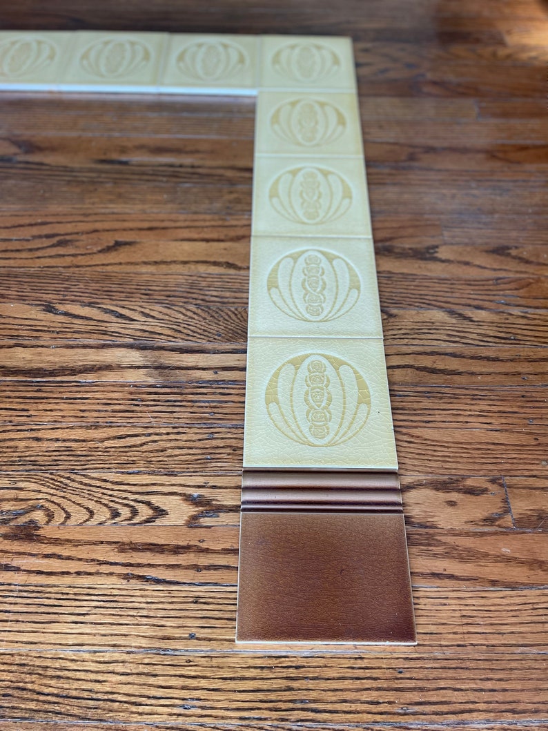 May include: A decorative tile border with a repeating pattern of stylized flowers in a light yellow color. The border is set against a dark brown wood floor.