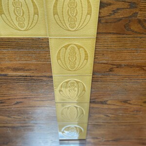 May include: A row of six yellow ceramic tiles with a repeating pattern of a stylized flower design. The tiles are arranged vertically and are set against a wood background.